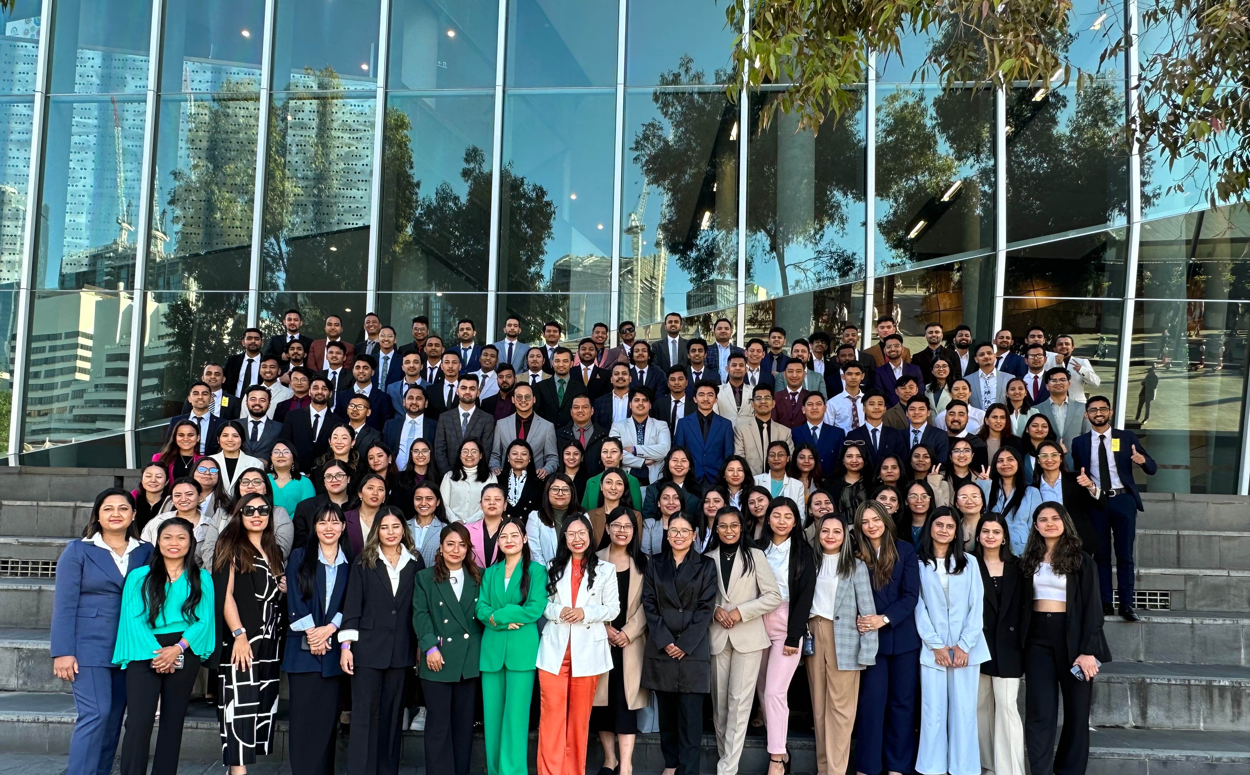 Large professional group photo of Stellars Group members in business attire standing in front of a modern glass building, showcasing the diverse and accomplished community of professionals and students